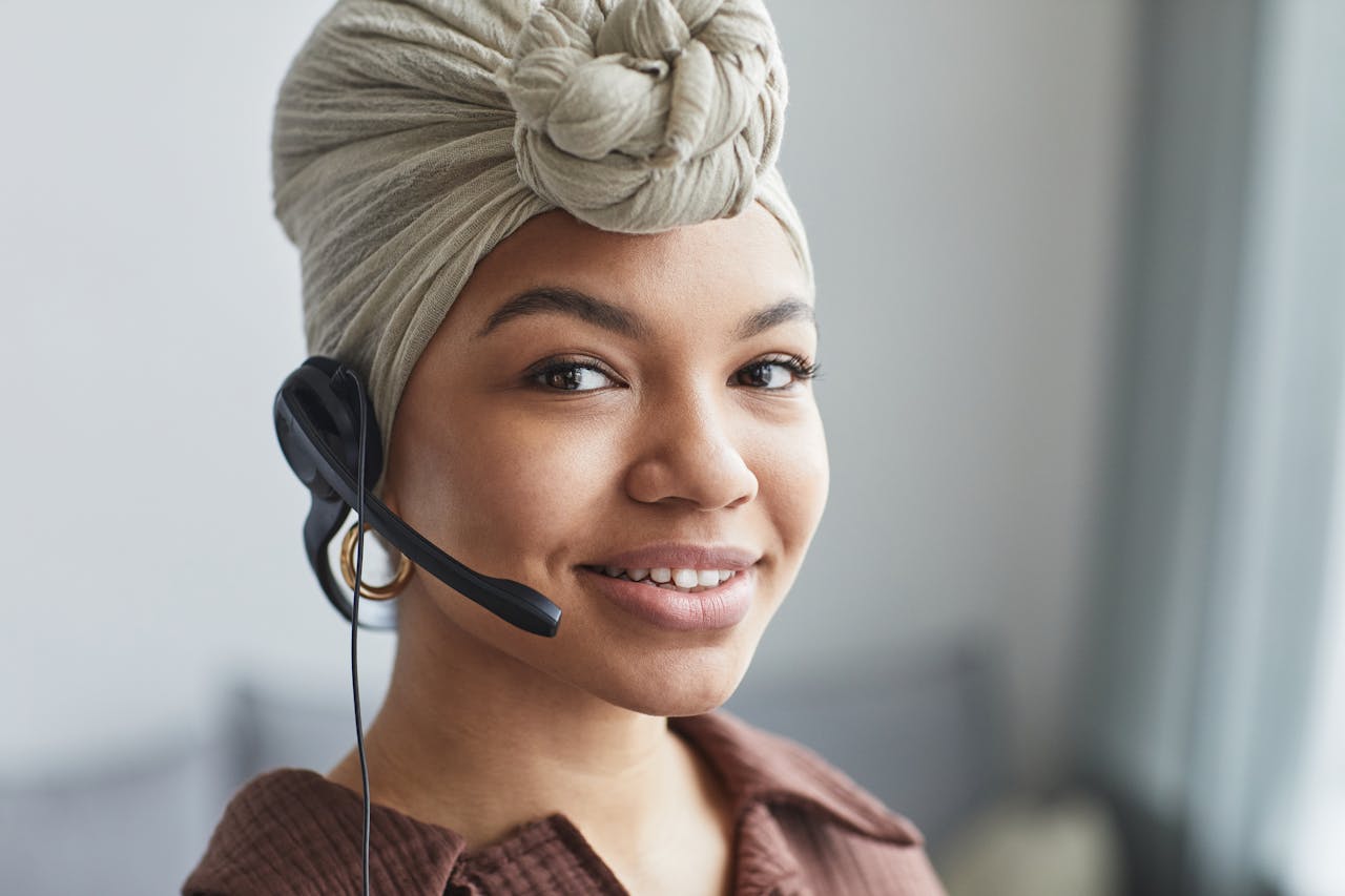 Delighted African American female call agent with headset wearing turban looking away while standing in light room on blurred background